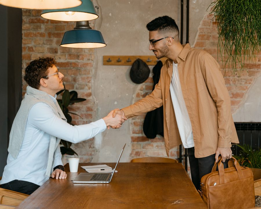Two people shaking hands across a desk at the start of a meeting, a laptop and notebook in front of them